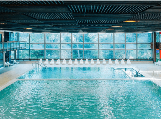 Piscina interna moderna con grandi finestre e sedie a sdraio bianche.